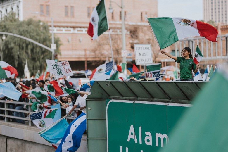 One person waves a Mexican flag while standing on the wax railing over a highway next to a Highway Street sign in downtown Los Angeles. In the background, demonstrators stop signs and flags on both sides that are set up along the railing.