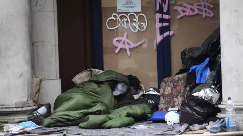 Pa Medien A Rauer sleep in a door, wrapped in a green sleeping bag, surrounded by their belongings and a pigeon