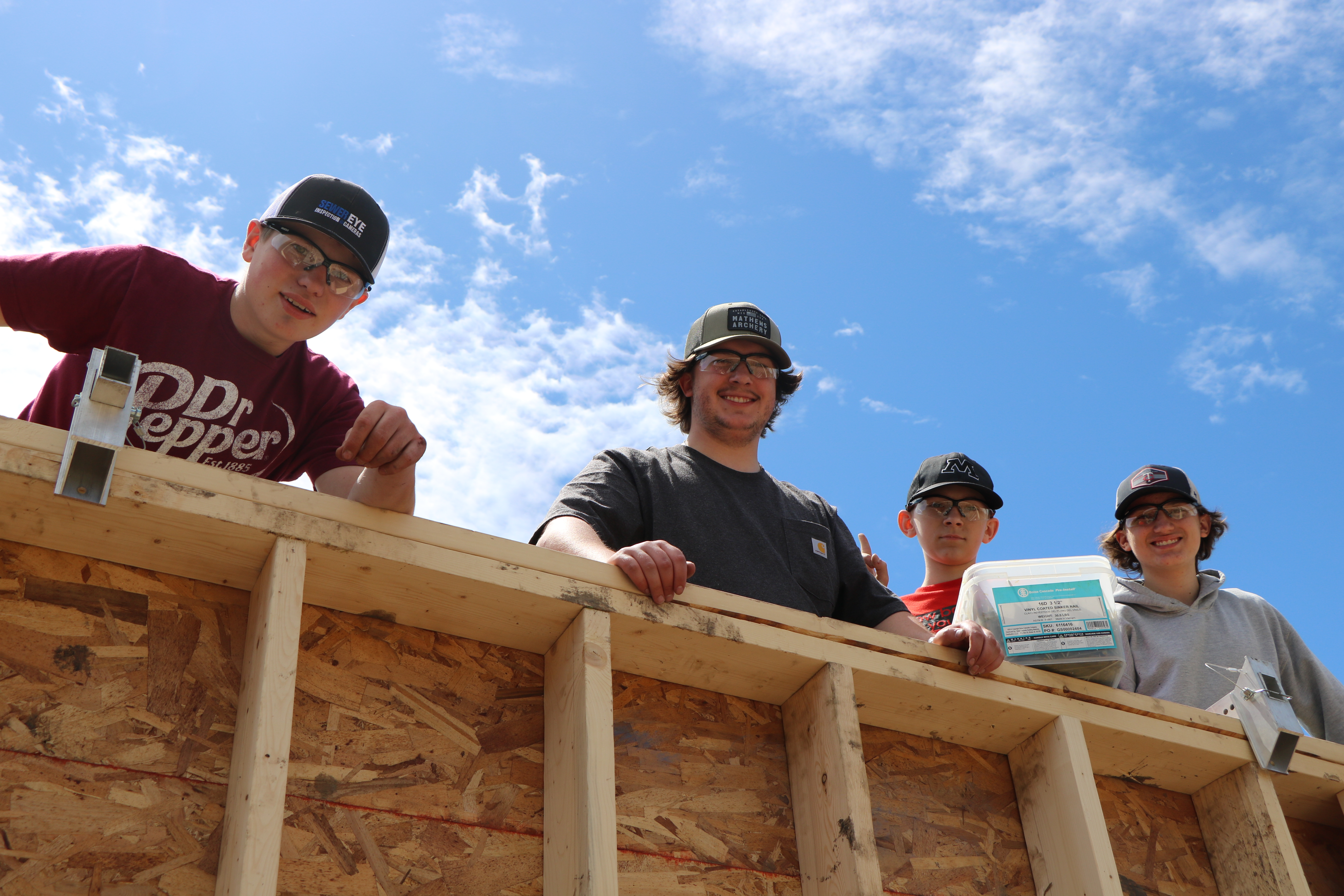 Four male teenagers who stand together in the camp of the Herdina Construction Trades 2024 look behind a under -groomed wall and smile in the camera.