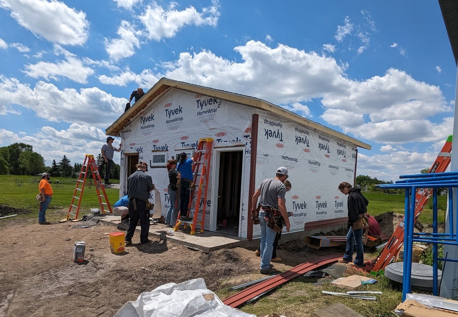 Several students and trainers in Camp 2024 Herdina Construction Trades are different jobs around the location of a small building from the construction.