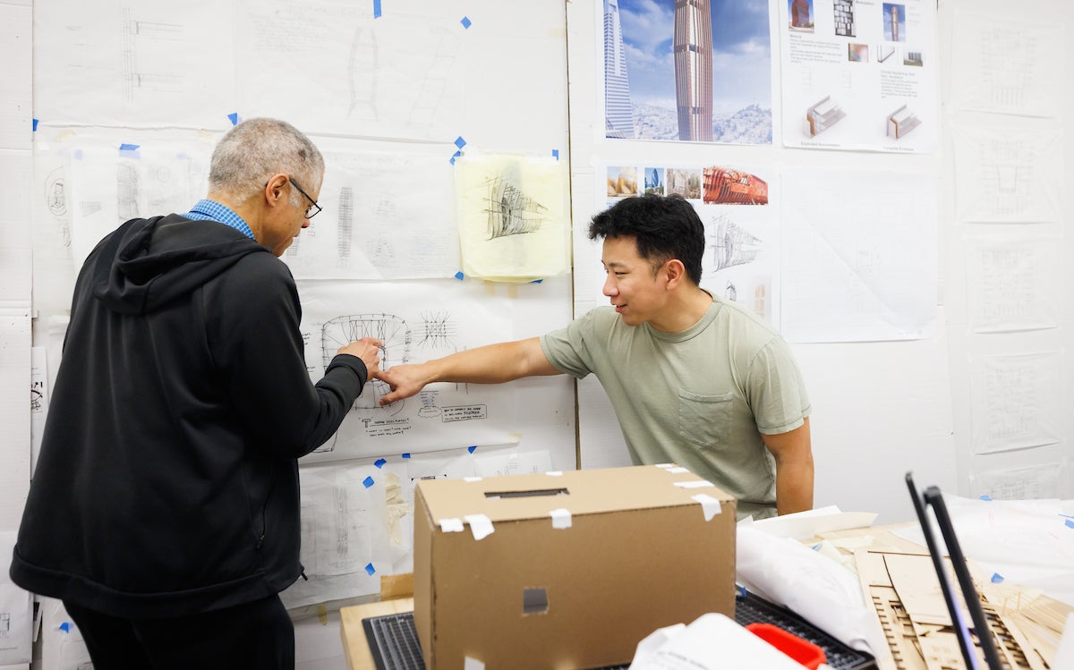 A professor and a student indicate a number of drawings on a wall of an Archtieur studio.