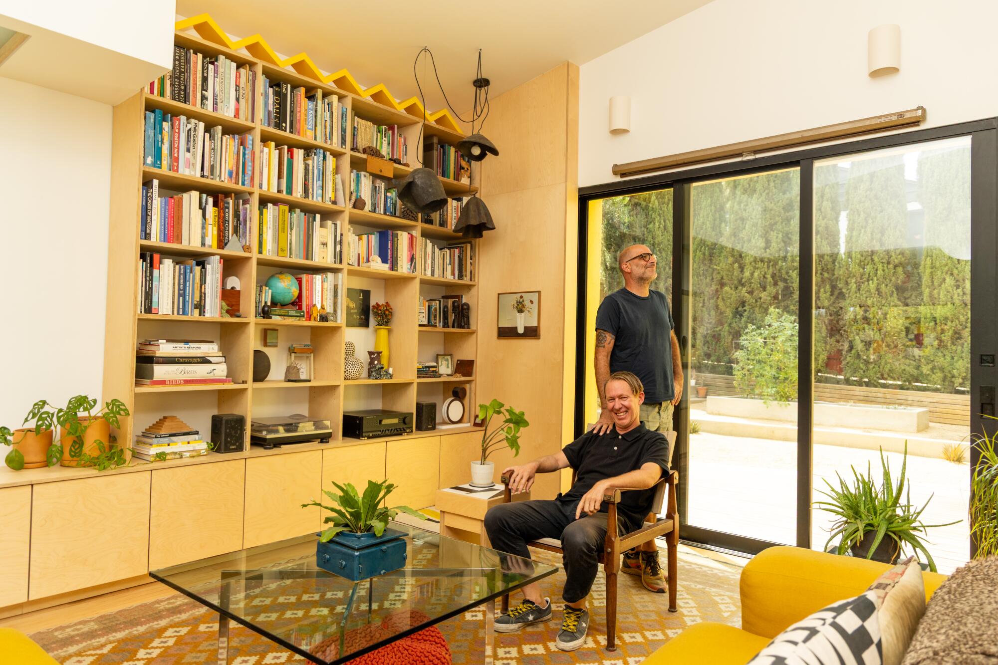 Two people, one sitting and the other standing, in a living room with bookshelves and drawers.