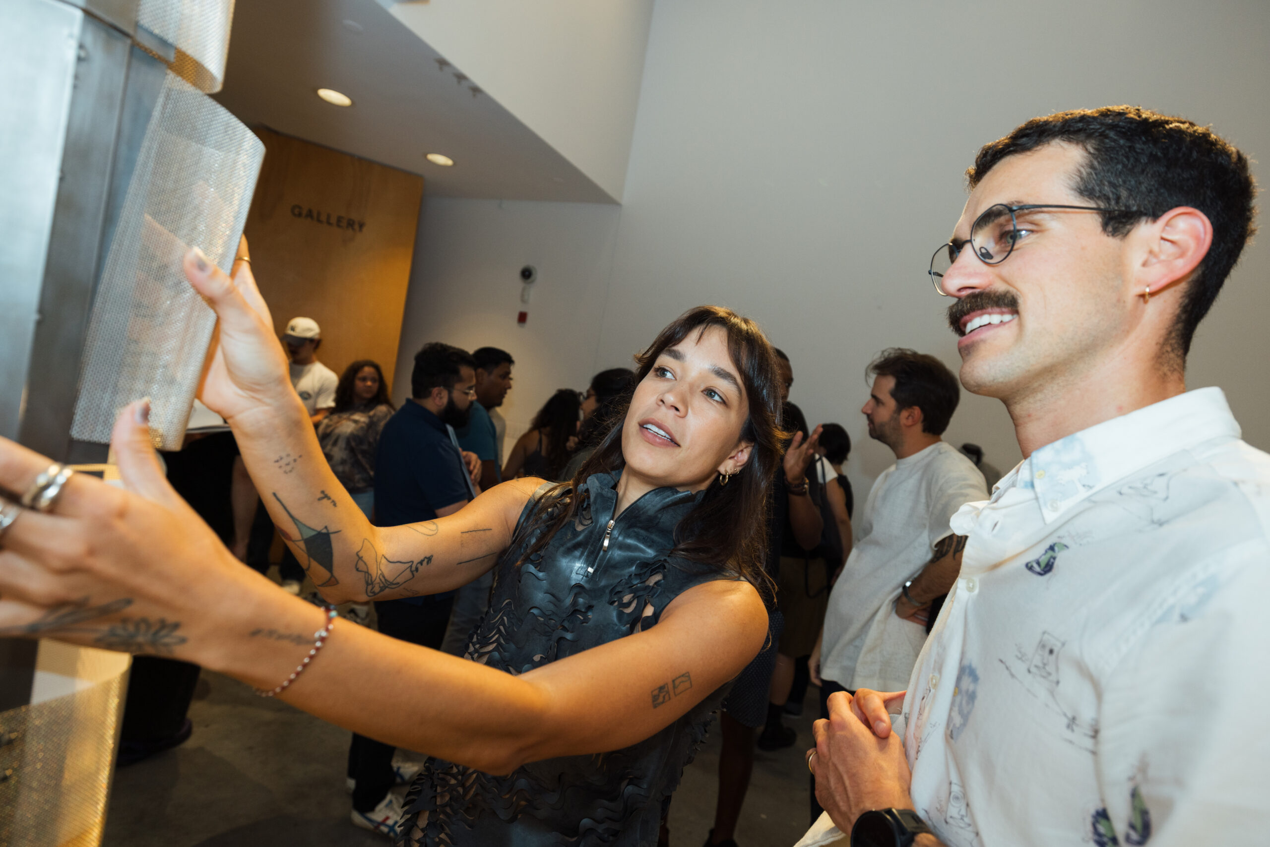 A woman with long hair and tattoos interacts with a metallic display and appears engaged and lively. She is wearing a sleeveless, structured outfit. A man with glasses and a mustache stands next to her, smiling and watching the interaction. Several other people can be seen in the background, contributing to a lively gallery atmosphere. The gallery sign is partially visible.
