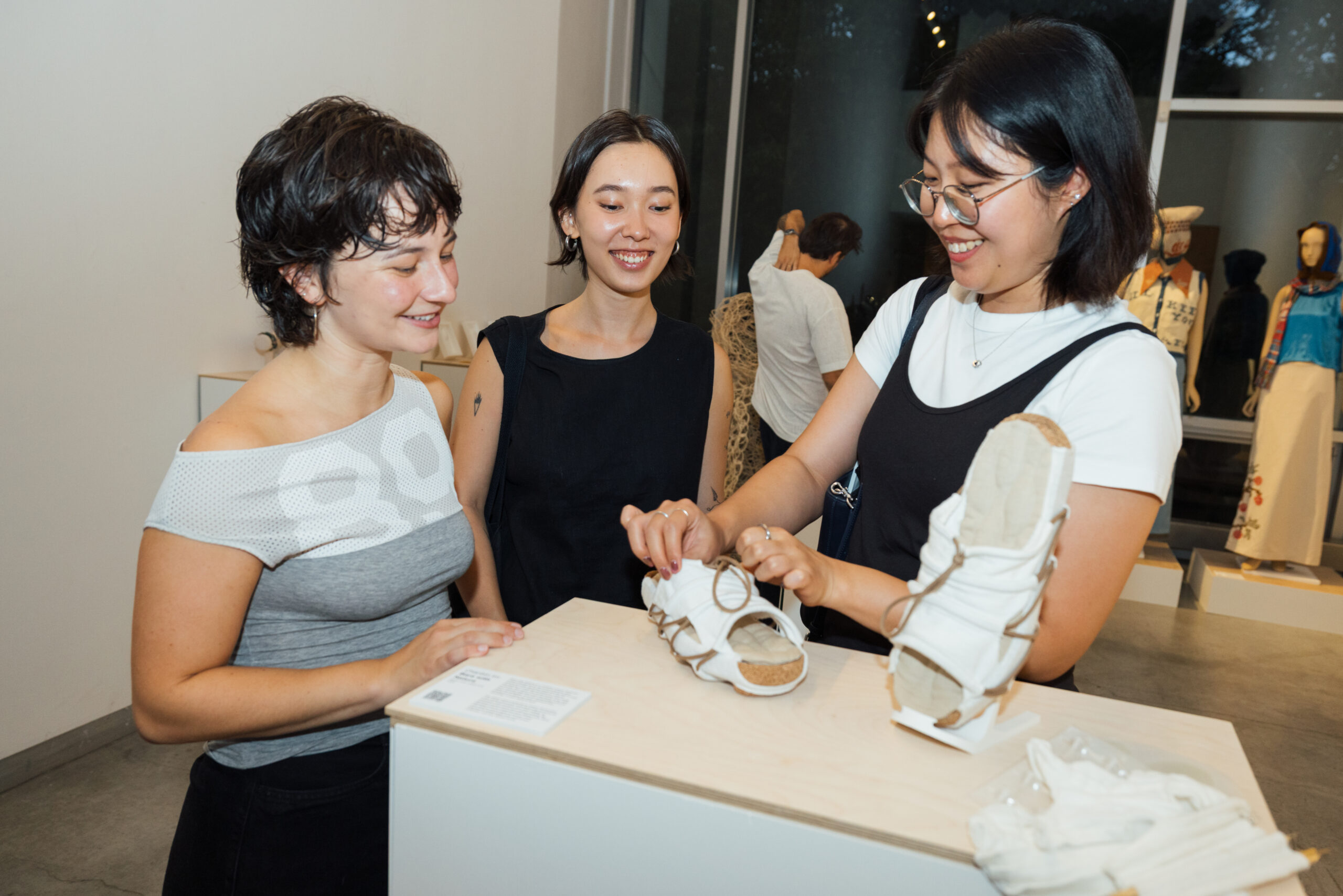 Three women talk at an art exhibition. A woman dressed in a gray off-the-shoulder top smiles as she leans against a display table. The second woman, wearing a black sleeveless dress, looks on with a smile. The third woman, wearing a black and white outfit, examines a pair of unique white sandals on the table. Other works of art can be seen in the background, including clothing displays.
