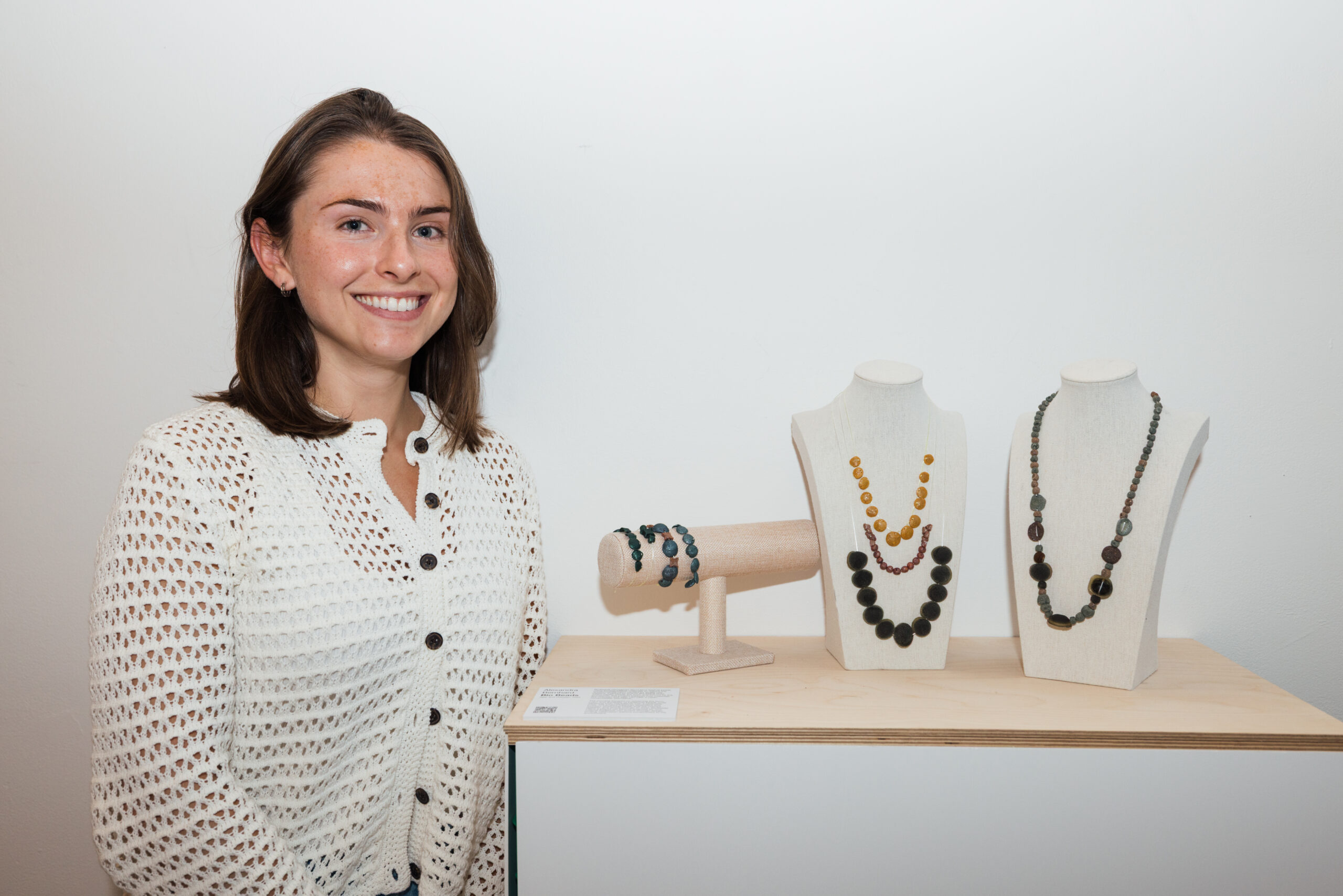 A smiling person stands next to a display of artistic sculptures in an exhibition room. They wear a dark, elaborately designed sleeveless top and light-colored pants. In the background, tall metal sculptures can be seen alongside smaller pieces mounted on stands, showcasing different materials and textures. A wooden door can partially be seen on the right.