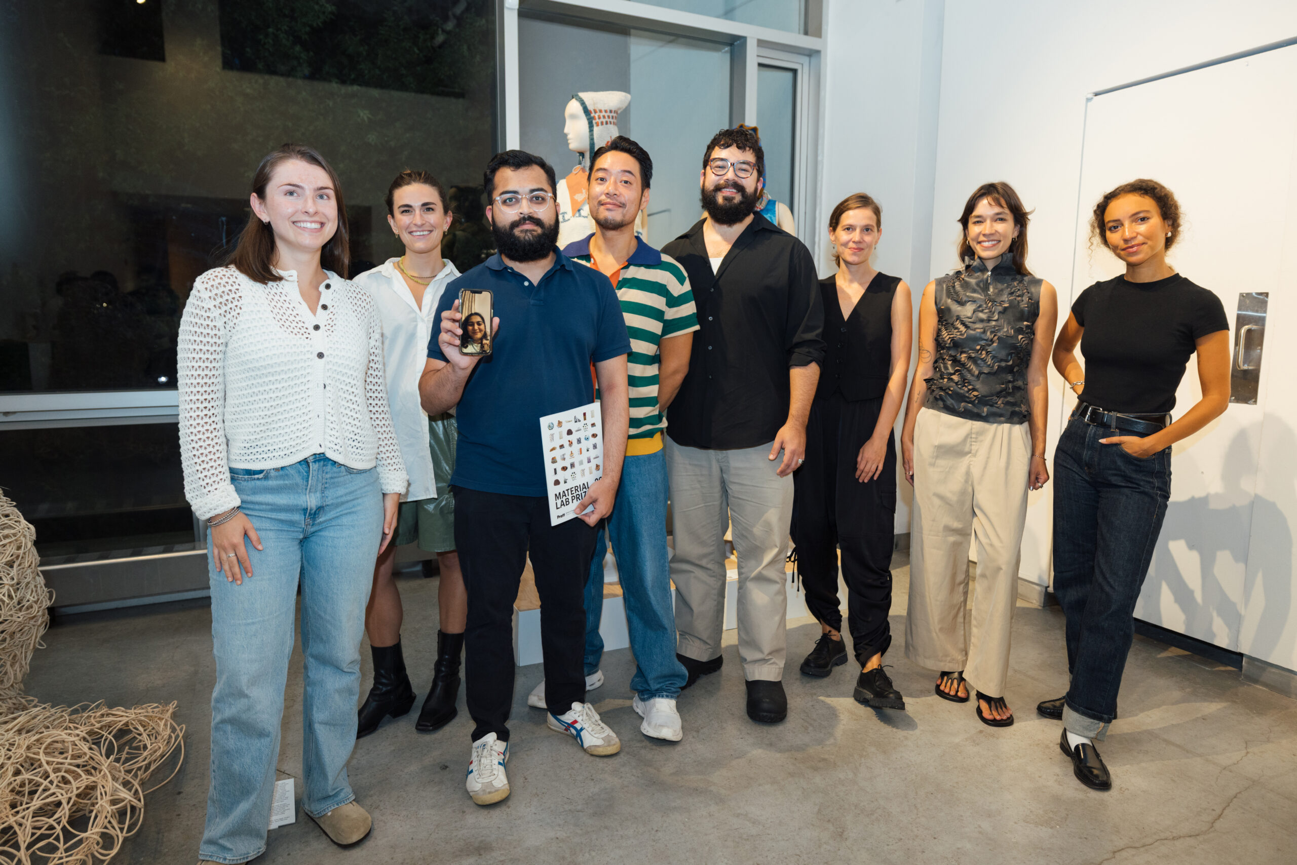 A group of eight people stand together in a well-lit interior and smile at the camera. They are dressed in various casual and stylish outfits. A person in the middle is holding a phone with a face on it, while another is holding an inscribed brochure "MATERIAL LABORATORY." A decorative mannequin can be seen behind it. The facility features modern furnishings with concrete floors and large windows.